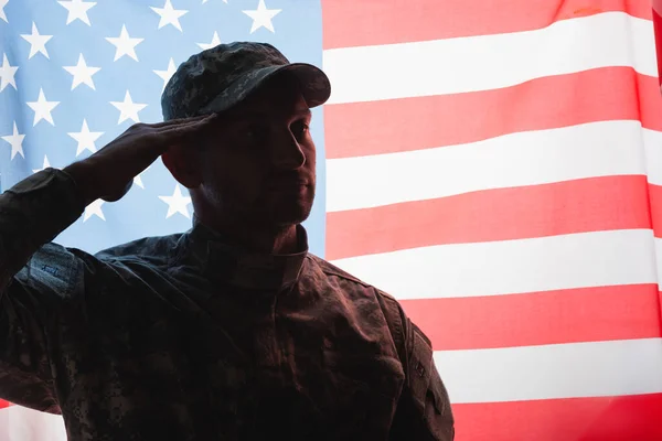 Patriotic military man in uniform and cap giving salute near american flag on background — Stock Photo