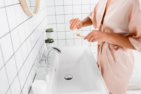 Обрізаний вид жінки в шовковому халаті, що наносить зубну пасту на зубну щітку у ванній — стокове фото