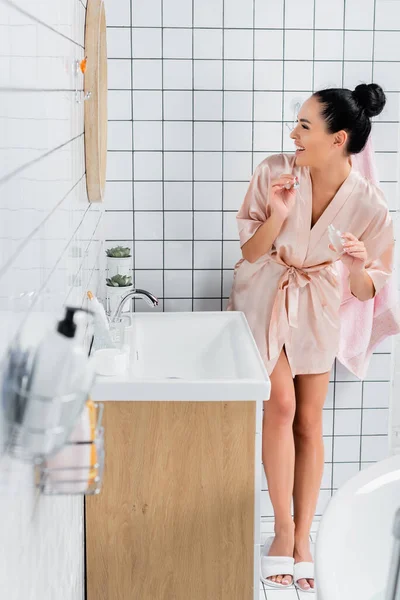 Mulher sorridente em roupão de banho de seda segurando soro cosmético no banheiro — Fotografia de Stock