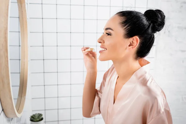 Mujer sonriente en albornoz de seda sosteniendo cepillo de dientes con pasta en el baño - foto de stock