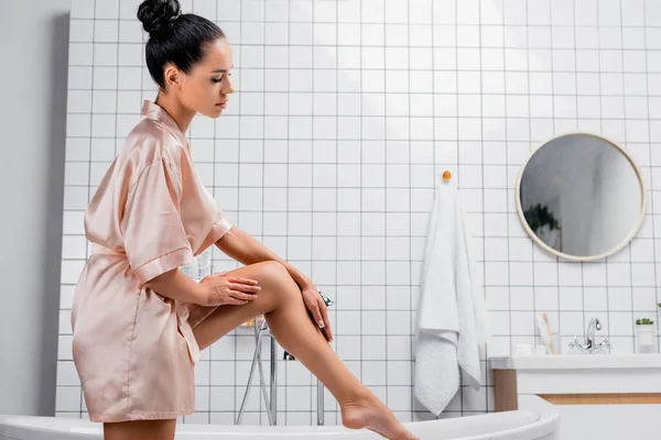 Vue latérale de la jeune femme en peignoir satiné touchant la jambe près de la baignoire — Photo de stock