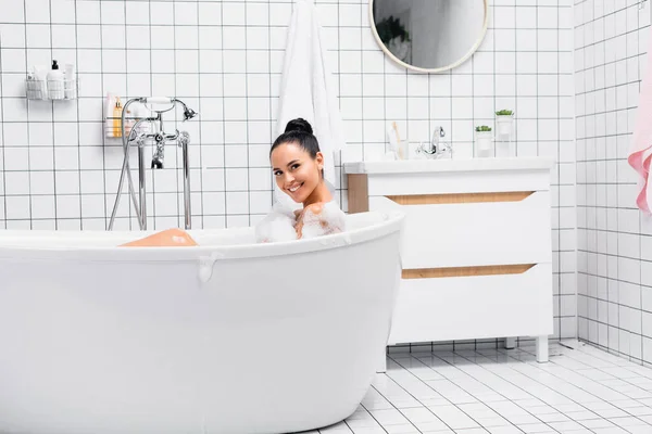 Cheerful brunette woman looking at camera while taking bath with foam — Stock Photo