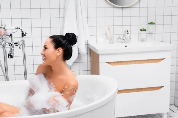 Mujer morena sonriendo mientras se sienta en la bañera con espuma - foto de stock