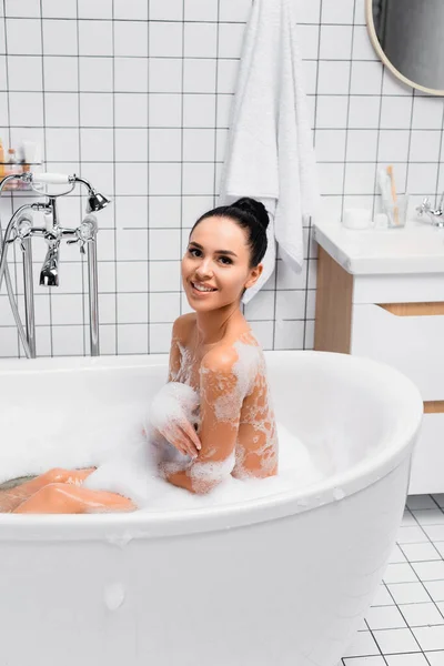 Bruna donna guardando la fotocamera mentre seduto nella vasca da bagno con schiuma in bagno moderno — Foto stock