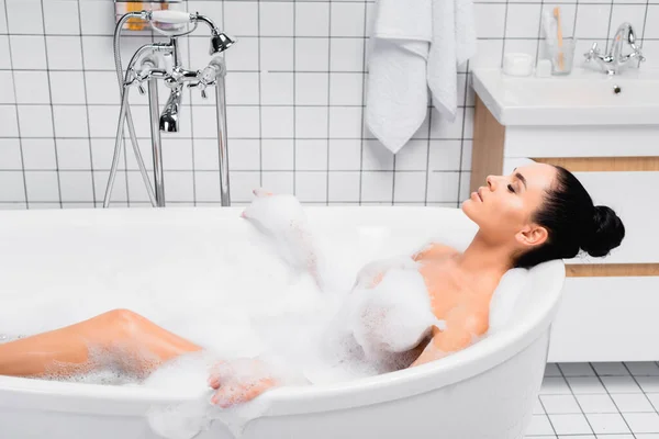 Brunette woman lying with closed eyes in bathtub with foam — Stock Photo
