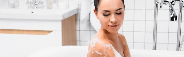 Young woman looking at wet shoulder in foam in bathtub, banner — Stock Photo