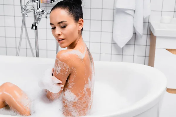 Mujer joven tocando el brazo en espuma mientras toma el baño en casa - foto de stock