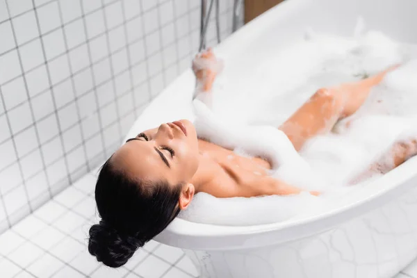 Brünette Frau entspannt sich in der Badewanne mit Schaum auf verschwommenem Hintergrund — Stockfoto