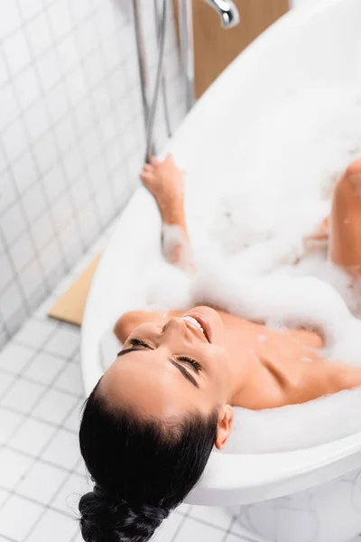 Vista ad alto angolo di donna sorridente che fa il bagno con schiuma su sfondo sfocato — Foto stock