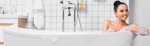 Donna attraente sorridente mentre si siede nella vasca da bagno con schiuma in bagno moderno, banner — Foto stock