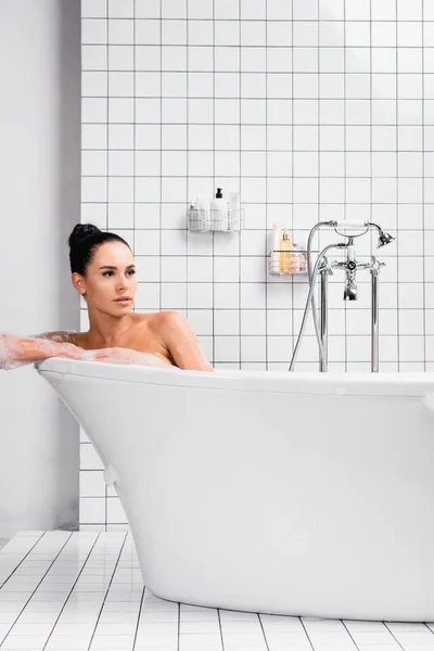 Brünette Frau schaut weg, während sie in der Nähe von Wasserhahn und Toilettenartikeln badet — Stockfoto