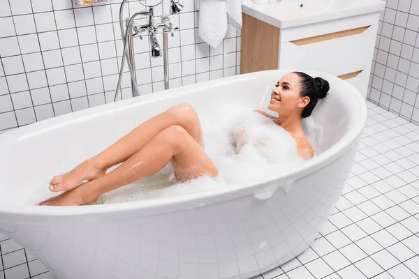 Jovem mulher sorrindo enquanto estava deitada na banheira com espuma em casa — Fotografia de Stock