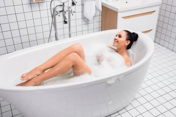 Jeune femme souriant tout en étant couché dans la baignoire avec mousse à la maison — Photo de stock