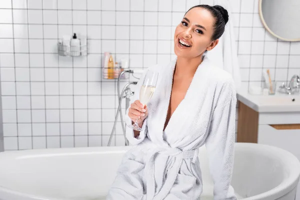 Smiling woman in bathrobe holding glass of champagne in bathroom — Stock Photo