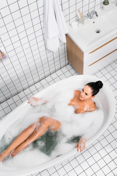 Vue aérienne de la femme sexy couchée dans le bain avec mousse près de la serviette dans la salle de bain — Photo de stock