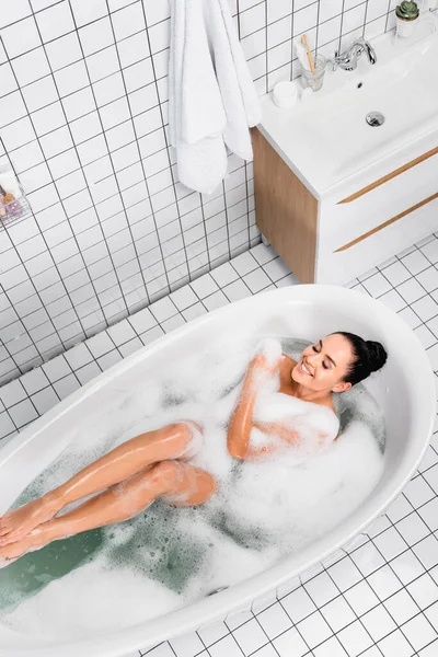 Draufsicht auf eine fröhliche Frau mit geschlossenen Augen, die in einer Badewanne mit Schaum genießt — Stockfoto