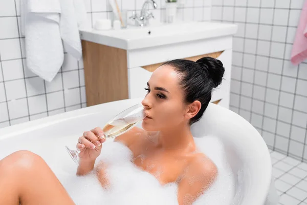 Mujer morena bebiendo champán en la bañera con espuma - foto de stock
