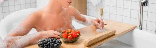 Cropped view of woman holding glass of champagne near fresh berries on bathtub tray, banner — Stock Photo
