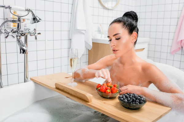 Femme brune prenant des fraises près d'un verre de champagne sur un plateau de baignoire — Photo de stock