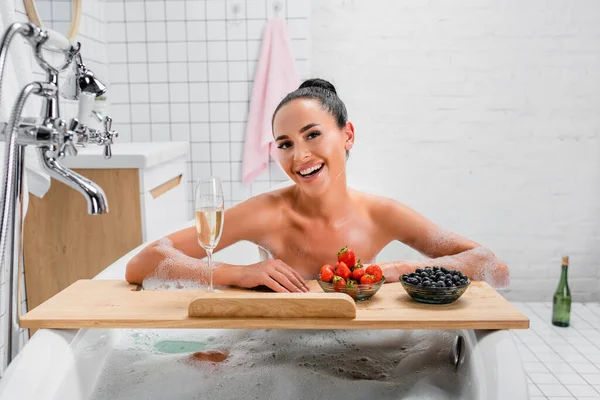 Donna allegra seduta nella vasca da bagno con schiuma vicino champagne e bacche sul vassoio — Foto stock