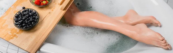 Vista recortada de bayas frescas en bandeja de madera cerca de la mujer tomando baño con espuma, pancarta - foto de stock