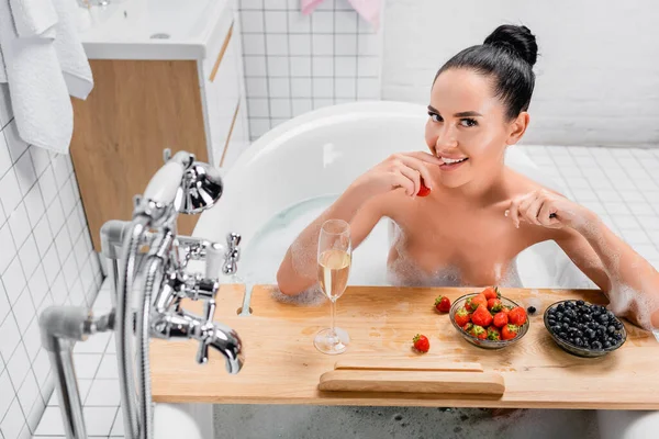 Femme sexy souriant tout en tenant fraise près du verre de champagne sur plateau en bois dans la baignoire avec mousse — Photo de stock