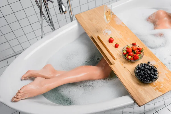 Cropped view of woman taking bath with foam near glass of champagne and fresh berries on tray — Stock Photo