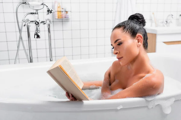 Чуттєва жінка читає книгу, сидячи у ванній з піною в сучасній ванній — стокове фото