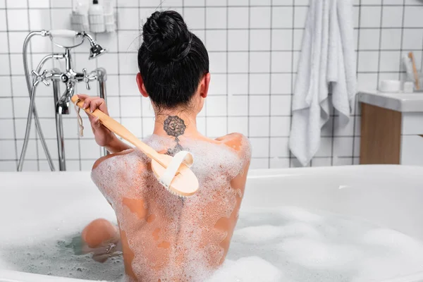 Vue arrière de la jeune femme utilisant une brosse dans une baignoire avec mousse — Photo de stock
