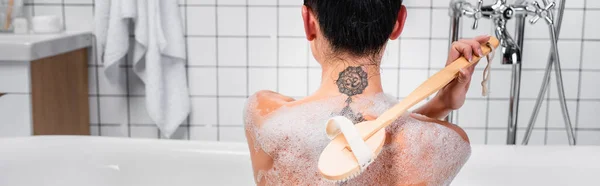 Vista posterior de la mujer tatuada usando cepillo en bañera con jabones, pancarta - foto de stock