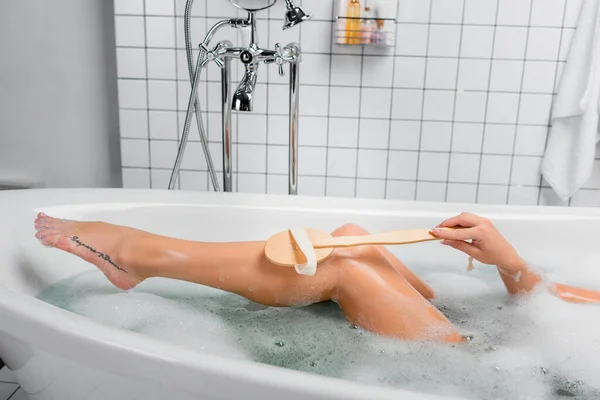 Vista recortada de una joven mujer limpiando la pierna con cepillo mientras toma el baño - foto de stock