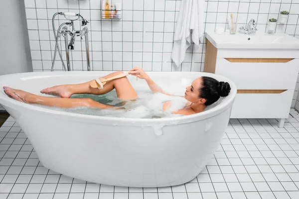 Mulher morena usando escova na banheira com espuma no banheiro moderno — Fotografia de Stock