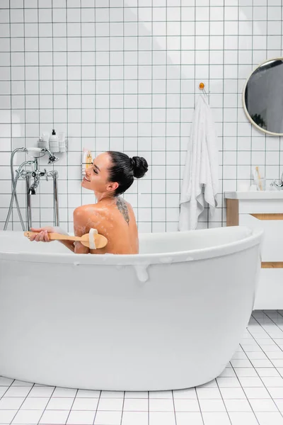 Fröhlich tätowierte Frau reinigt Körper mit Bürste beim Baden — Stockfoto