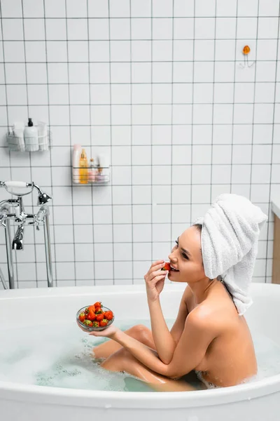 Mujer sexy con toalla en la cabeza comiendo fresa fresca mientras toma baño - foto de stock