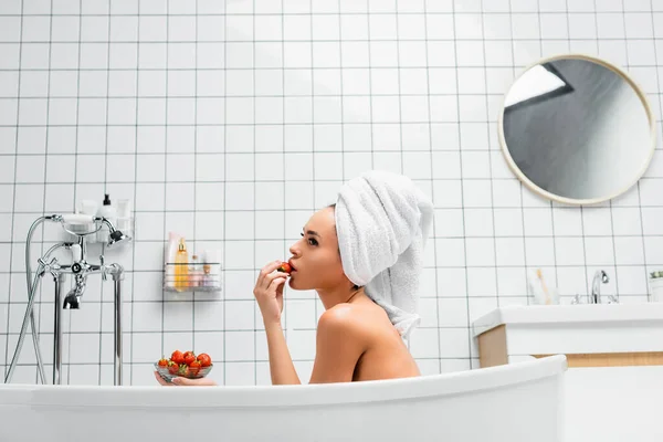 Side view of young woman with towel on head eating juicy strawberry in bathtub at home — Stock Photo