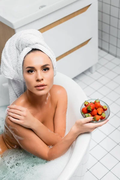 Молода жінка з рушником на голові дивиться на камеру, тримаючи миску полуниці у ванній — стокове фото