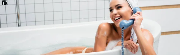 Mujer sonriente con toalla en la cabeza hablando por teléfono en baño con espuma, pancarta - foto de stock