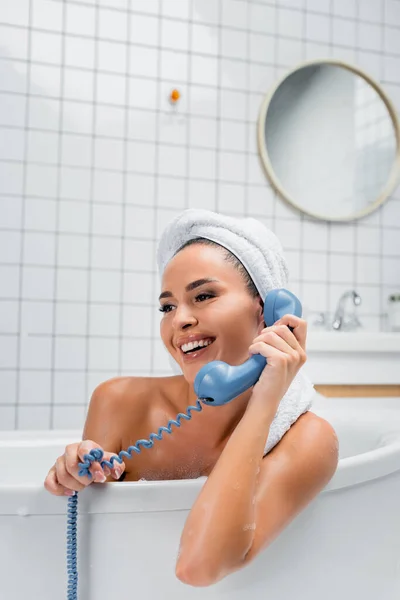 Cheerful woman with towel on head talking on vintage telephone in bath — Stock Photo
