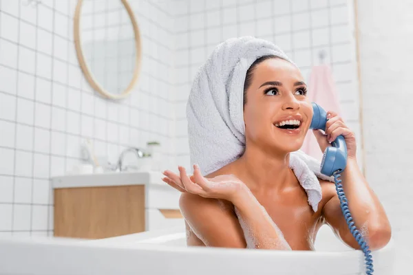 Mujer alegre en espuma y toalla en la cabeza hablando por teléfono en la bañera en primer plano borrosa - foto de stock