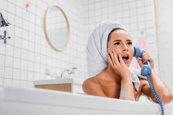 Femme choquée en serviette parlant au téléphone alors qu'elle était assise dans la baignoire à la maison — Photo de stock