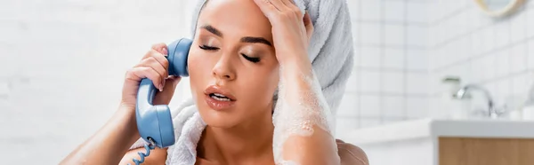 Femme fatiguée en mousse et serviette sur la tête parlant au téléphone dans la salle de bain sur fond flou, bannière — Photo de stock