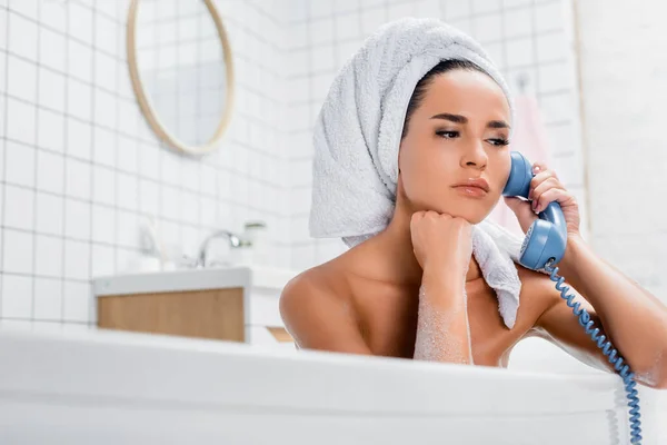 Donna pensierosa in asciugamano sulla testa che parla al telefono blu nella vasca da bagno in primo piano sfocato — Foto stock