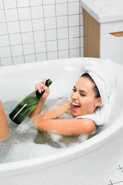 Donna eccitata con asciugamano in testa bottiglia di champagne in bagno con schiuma — Stock Photo