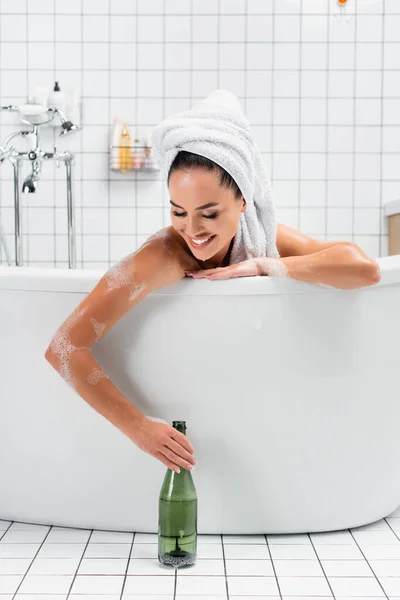 Mulher sorridente com toalha na cabeça tomando garrafa de champanhe enquanto toma banho — Fotografia de Stock