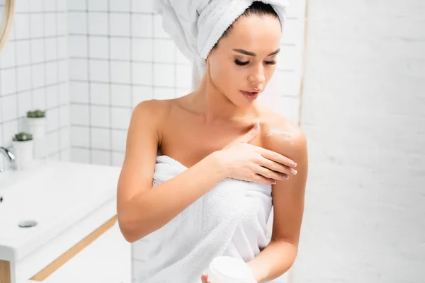 Jovem mulher em toalhas aplicando creme cosmético no ombro no banheiro — Fotografia de Stock