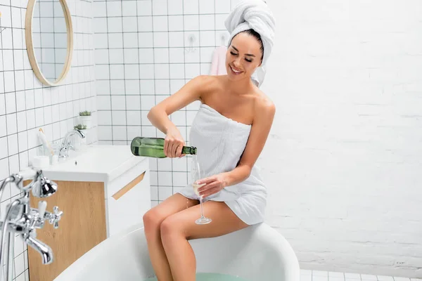 Cheerful woman in towels pouring champagne in glass near bathtub at home — Stock Photo