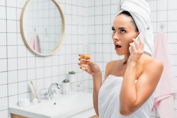 Geschockte Frau in Handtüchern spricht auf Smartphone und hält Glas Champagner im Badezimmer vor verschwommenem Hintergrund — Stockfoto