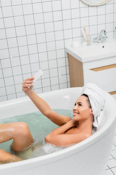 Mujer joven con toalla en la cabeza tomando selfie con teléfono inteligente mientras toma el baño - foto de stock