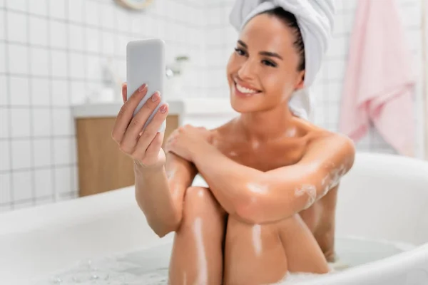 Smartphone in mano di donna sorridente in asciugamano prendere selfie in bagno su sfondo sfocato — Foto stock