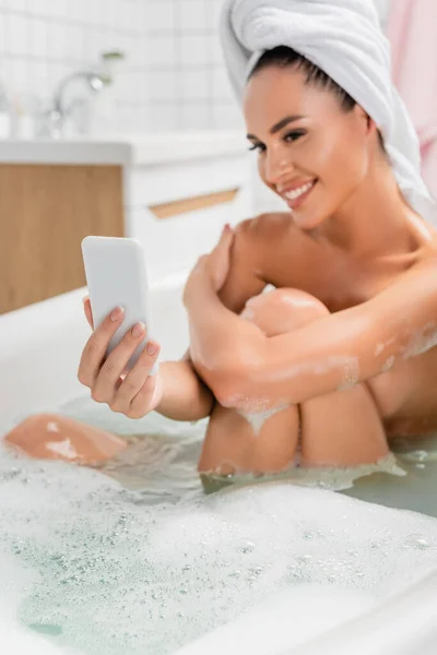Smartphone en la mano de la mujer joven en la toalla tomando selfie en el teléfono inteligente sobre fondo borroso en el baño - foto de stock